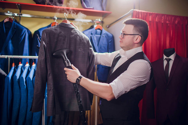 tailor cuts and sews a suit. A young man in the tailoring shop makes mens clothing. Custom-made clothing workshop. holds in her hand an electric iron. wet-heat treatment - Фото, изображение