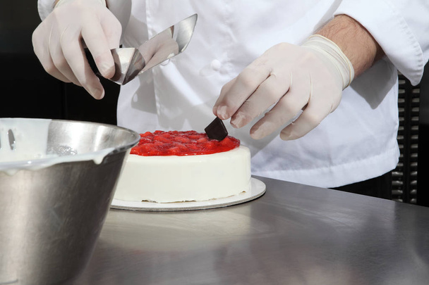 hands pastry chef prepares a cake, cover with icing and decorate with strawberries, works on a stainless steel industrial kitchen work top - Photo, image