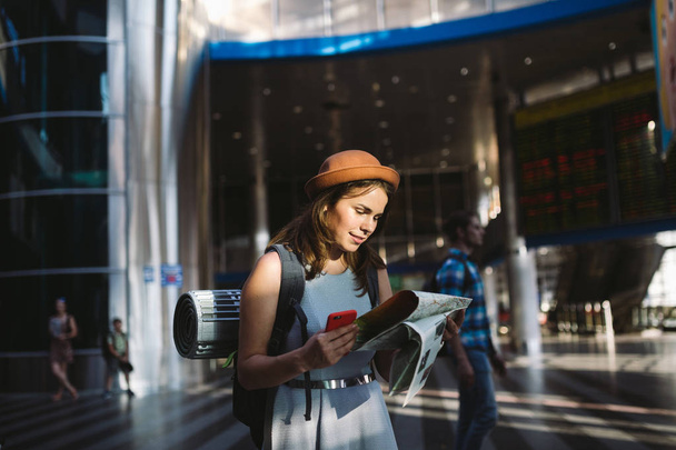 тематические путешествия и транспорт. Красивая молодая белая женщина в платье и рюкзак, стоящий в терминале железнодорожного вокзала, глядя на электронное табло с телефоном, карты бумаги ручной навигации
. - Фото, изображение