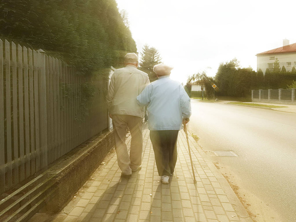 Ένα ζευγάρι των ηλικιωμένων με τα πόδια κατά μήκος του πεζοδρομίου κατά μήκος του δρόμου κρατώντας τα χέρια. Παππού και της γιαγιάς σε έναν περίπατο σε μια κατοικημένη περιοχή. Κίνημα υγεία των ηλικιωμένων. Ευτυχισμένη τρίτη ηλικία - Φωτογραφία, εικόνα