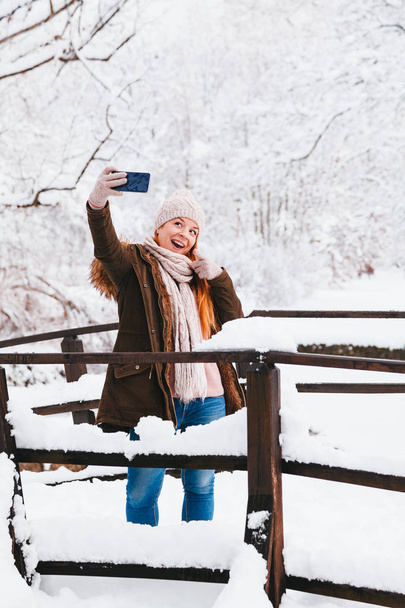 Nuori nainen ottaa selfien puistossa lumisena talvipäivänä
 - Valokuva, kuva