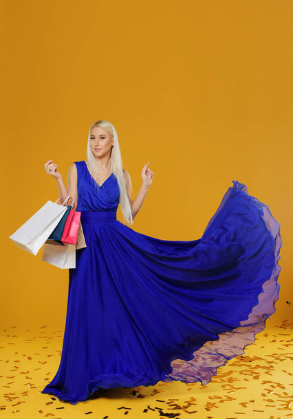 Blonde woman in a  blue luxury dress with bag on black friday shopping. Copy space for sale ads. Yellow background - Φωτογραφία, εικόνα