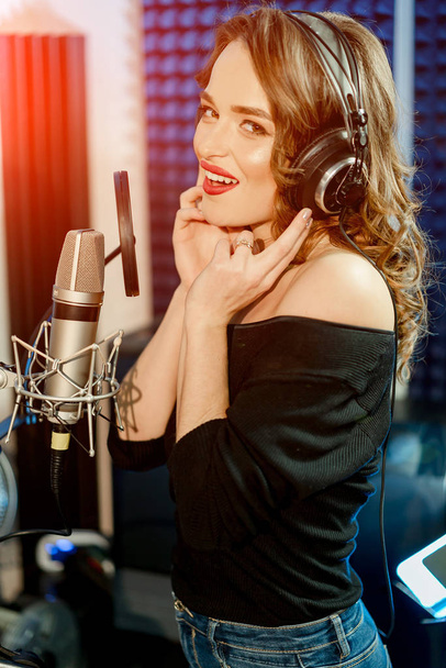 Shot of a lovey young model in headphones singing with a microphone and looking at camera in a professional studio. Beautiful young woman with microphone and headphones. - Foto, Imagen