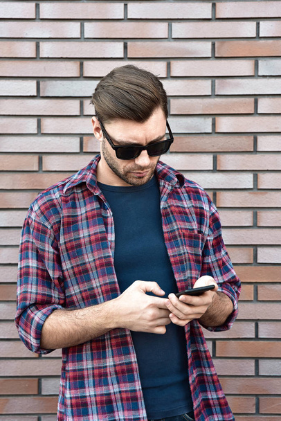 Vista frontal del joven guapo en ropa casual inteligente sostener un teléfono móvil mientras está de pie en el fondo de la pared de ladrillo
. - Foto, imagen