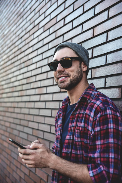 Hipster sms texting phone app in city street on brick wall background. Amazing man holding smartphone in smart casual wear standing. Urban young professional lifestyle. - Photo, Image