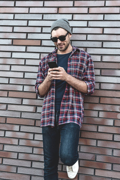 Додаток для смс-повідомлень Hipster на вулиці міста на фоні цегляної стіни. Дивовижний чоловік тримає смартфон у розумному повсякденному одязі. Міський молодий професійний спосіб життя
. - Фото, зображення