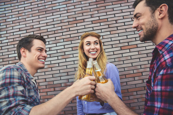Троє щасливих друзів насолоджуються пивом на фоні коричневої цегляної стіни
. - Фото, зображення