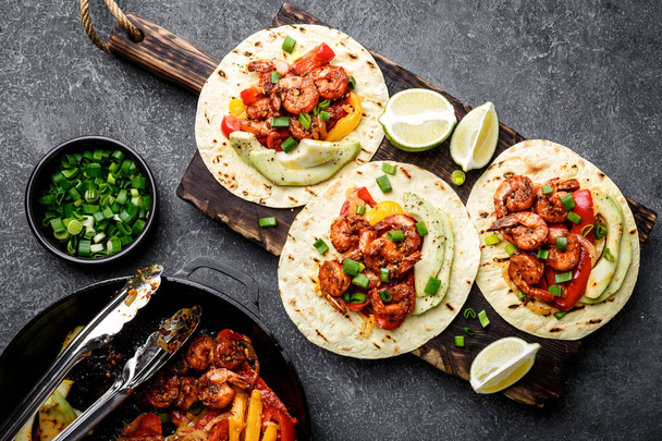 Fajitas in tortillas with fried shrimps, bell peppers and onion served up with avocado and green onions on wooden cutting board, top view, flat lay - Photo, Image