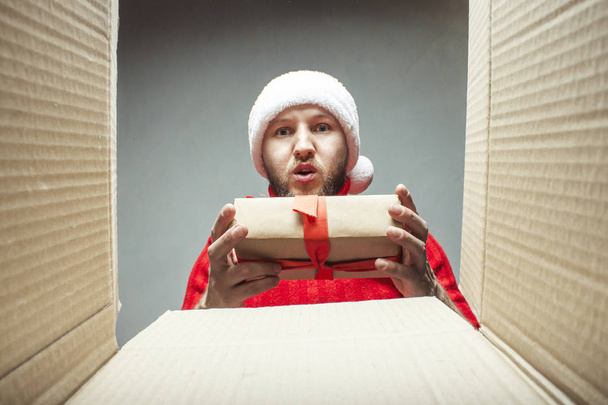 Mann mit Weihnachtsmannmütze und rotem Pullover legt sich hin, nimmt Geschenke ein oder aus der Kiste. Konzept der Verpackung und des Versands oder Empfangens von Geschenken für Freunde und Verwandte zu Weihnachten, Neujahr - Foto, Bild