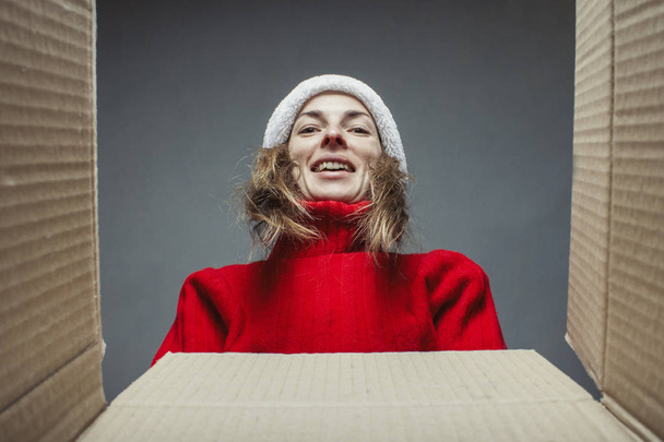 Girl in the hat of Santa Claus and a red sweater looks at the contents of the box. Concept of opening or sending gifts, surprise, package, shopping on Internet for Christmas, New Year - Fotografie, Obrázek