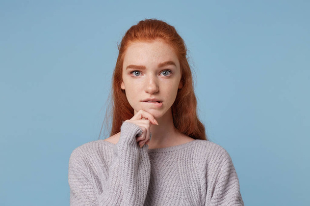 Portrait of a girl with red hair, looks camera holds her hand near the chin bit her lip, reflects on something, feels a little hesitant wants to make some decision, doubts, isolated on blue background - Photo, image