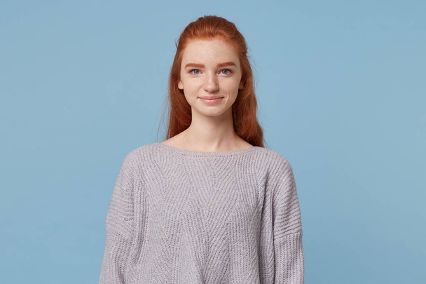 Jonge aantrekkelijke mooi vrij roodharige meisje met lang haar ziet er in de camera, glimlach zoet, is gekleed in een oversized eenvoudige warme trui, geïsoleerd op een blauwe achtergrond - Foto, afbeelding