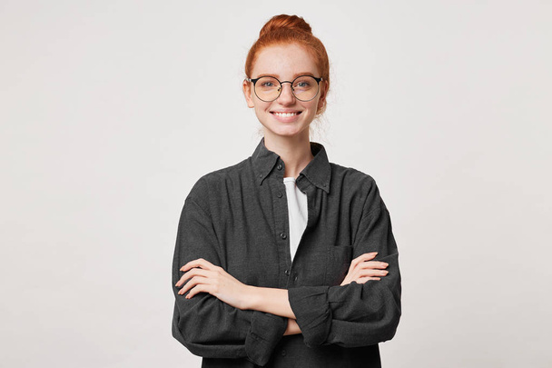 Self-confident red-haired girl with hair gathered in bun in a men's black shirt, glasses stands smiling with arms crossed - Valokuva, kuva