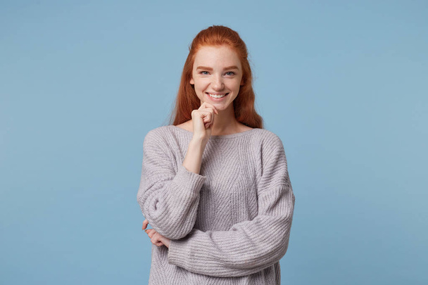 Een vrolijke openhartig vrij aantrekkelijk meisje met rood haar staat breed glimlachend, in een warme oversized trui met haar hand in de buurt van haar kin, over blauwe achtergrond - Foto, afbeelding