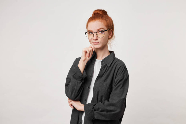 Portrait of a young red-haired girl in glasses who looks pensively to the side, dressed in a black men's shirt, keeps her hand on her chin, doubts about some solution, ponders the problem - Fotó, kép
