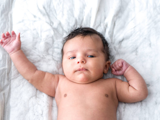 Portrait of a mixed race handsome and cute naked baby boy with lots of brown hair flexing his muscles with one arm and waving with the other laying on his back on a white pad background. - Photo, Image