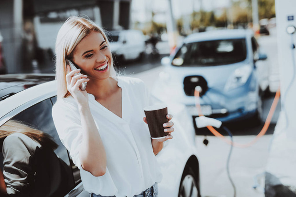 Tesla Car Charge Concept. Girl Talks The Phone. Automobile Station. Innovation Technology. New Generation Electro Hybrid Vehicle Plugin. Ecology Station. Futuristic Power. Cup Of Coffee. - Foto, imagen