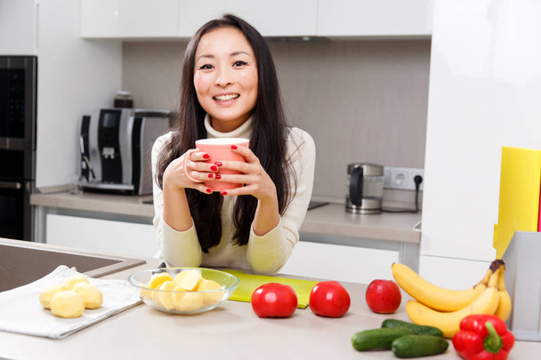 Image of happy woman with mug in hands standing at table with vegetables and fruits - Фото, зображення