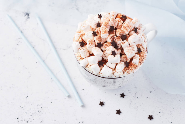 Hot chocolate or cocoa with whipped cream and marshmallow candy sprinkled with cinnamon or cocoa powder with chocolate stars on white marble table and blue silk in the background. Copy space. - 写真・画像