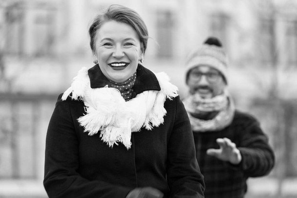 Stylish charming middle aged woman looking at you and expressing happiness while her positive husband making attempts for surprising her on the background - Фото, зображення