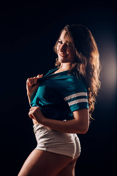 Portrait of sexy attractive hot brunette woman with long wavy hair in short white shorts and blue t-shirt in the dark on black background. Slim figure, fitness, sports, dancing, twerk, hobby concepts - Photo, image