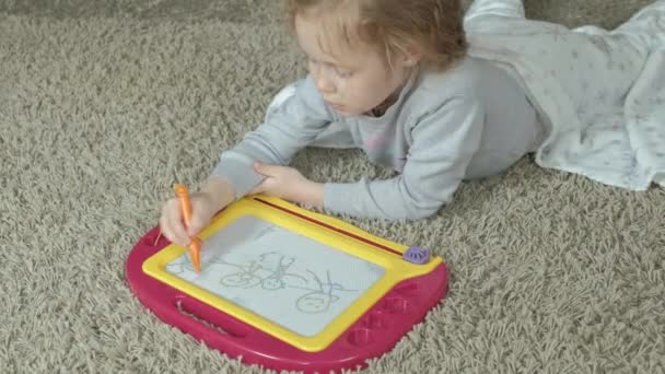 Une petite fille aux cheveux roux ondulés se couche sur le sol et dessine sur un tableau magnétique. Le concept du processus éducatif
. - Séquence, vidéo