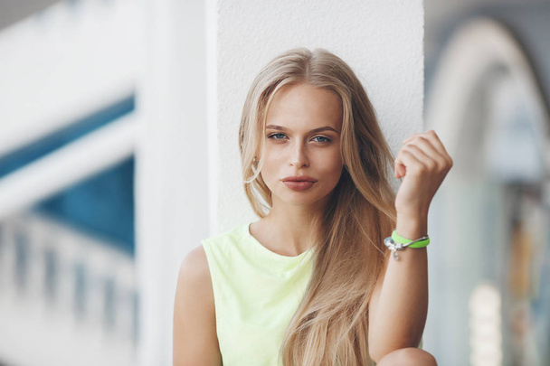Photo of a beautiful blonde girl sitting on the railing in the hotel at sunset. Woman resting on the street and waiting for friends after a work shift.Cute blue-eyed blonde poses alone sitting on a white railing on the hotels outdoor terrace - Photo, Image