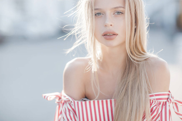 A young beautiful woman with long, straight hair and gray eyes, beautiful makeup, pink plump lips, sweet smile, blonde, spending her time outdoors in the summer in the city, dressed in a white jersey and checkered shirt, posing in the sunlight - 写真・画像