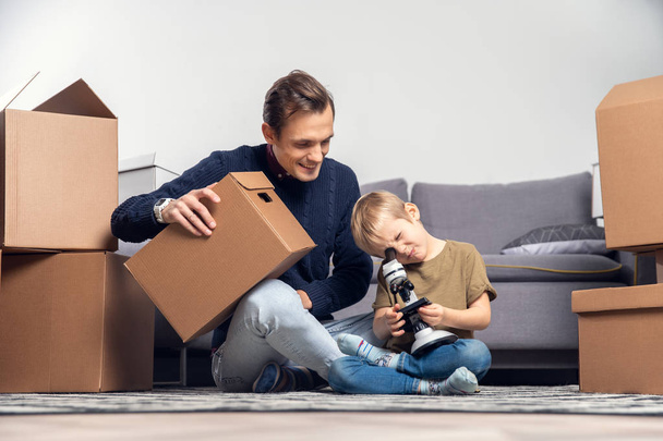 Picture of happy man and small boy with microscope sitting on floor among cardboard boxes in room - Fotó, kép