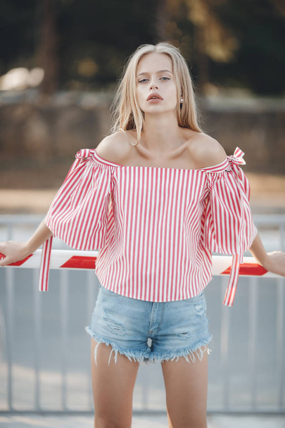 A young beautiful woman with long, straight hair and gray eyes, beautiful makeup, pink plump lips, sweet smile, blonde, spending her time outdoors in the summer in the city, dressed in a white jersey and checkered shirt, posing in the sunlight - Fotografie, Obrázek