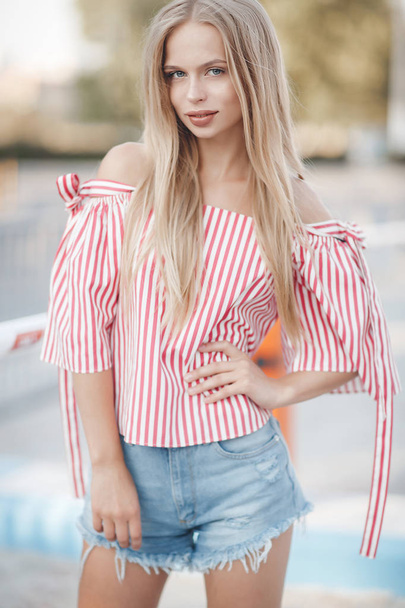 A young beautiful woman with long, straight hair and gray eyes, beautiful makeup, pink plump lips, sweet smile, blonde, spending her time outdoors in the summer in the city, dressed in a white jersey and checkered shirt, posing in the sunlight - Фото, изображение