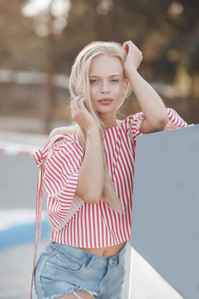 A young beautiful woman with long, straight hair and gray eyes, beautiful makeup, pink plump lips, sweet smile, blonde, spending her time outdoors in the summer in the city, dressed in a white jersey and checkered shirt, posing in the sunlight - Φωτογραφία, εικόνα