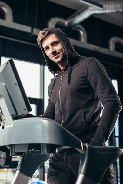 yakışıklı sporcu spor salonunda treadmill egzersiz ve kameraya bakarak hoodie - Fotoğraf, Görsel