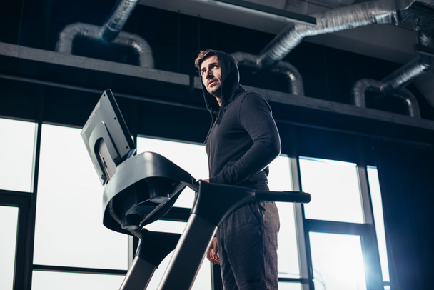 low angle view of handsome sportsman in hoodie exercising on treadmill in gym - Photo, Image