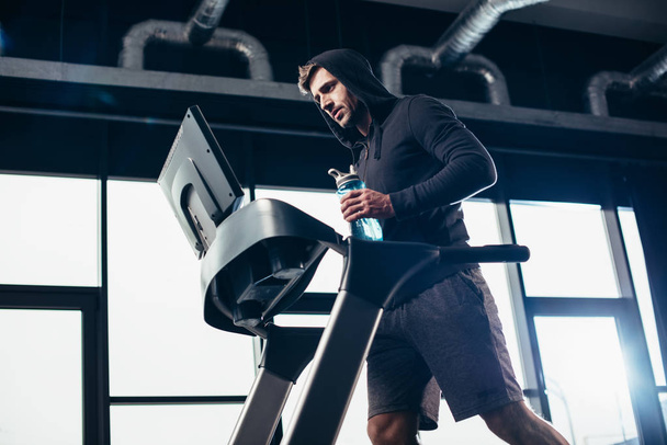 χαμηλή γωνία θέα όμορφος αθλητικός τύπος σε hoodie άσκηση σε διάδρομο και κρατώντας αθλητισμό μπουκάλι με νερό στο γυμναστήριο - Φωτογραφία, εικόνα