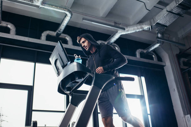 низкий угол зрения красивый спортивный мужчина в толстовке упражнения на беговой дорожке в тренажерном зале
 - Фото, изображение