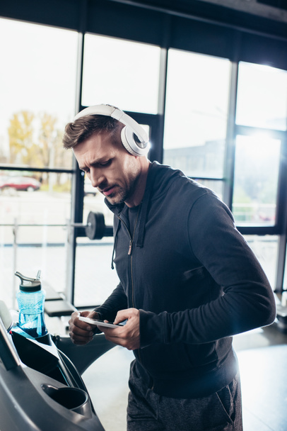 όμορφος αθλητικός τύπος σε hoodie άσκηση στο διάδρομο και χρησιμοποιώντας το smartphone στο γυμναστήριο  - Φωτογραφία, εικόνα