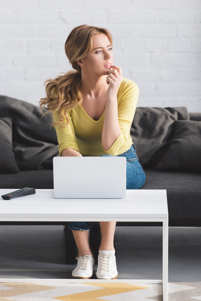 thoughtful woman looking away while sitting on couch and using laptop - Photo, Image