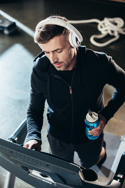 high angle view of handsome sportsman in hoodie exercising on treadmill and holding sport bottle in gym  - Фото, зображення