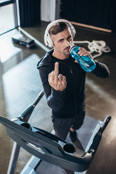 high angle view of handsome sportsman in hoodie showing middle finger on treadmill in gym  - Fotografie, Obrázek