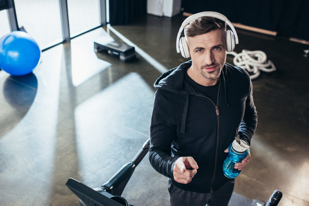 high angle view of handsome sportsman in hoodie standing on treadmill with sport bottle in gym  - Фото, зображення