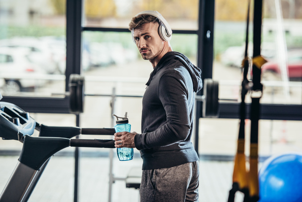 portrait of handsome sportsman in hoodie standing near treadmill with sport bottle in gym and looking at camera - Valokuva, kuva