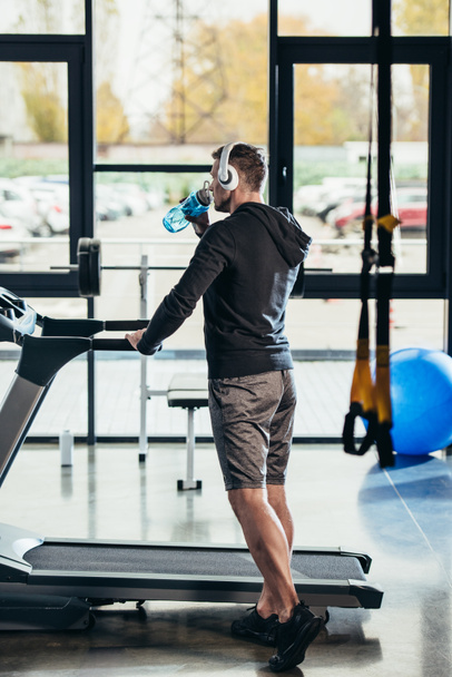 back view of sportsman in hoodie standing near treadmill with sport bottle in gym  - Fotó, kép