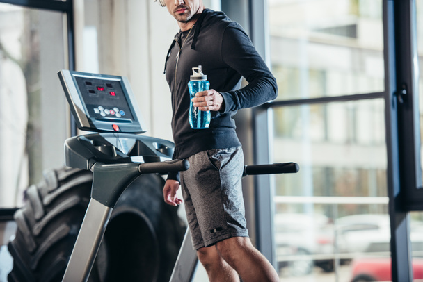 cropped image of sportsman in hoodie exercising on treadmill and holding sport bottle in gym  - Фото, изображение