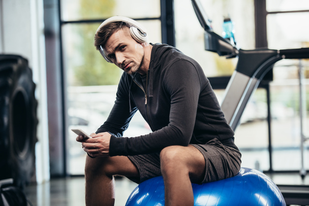 apuesto deportista sentado en la pelota de fitness, sosteniendo el teléfono inteligente y mirando a la cámara en el gimnasio
  - Foto, Imagen