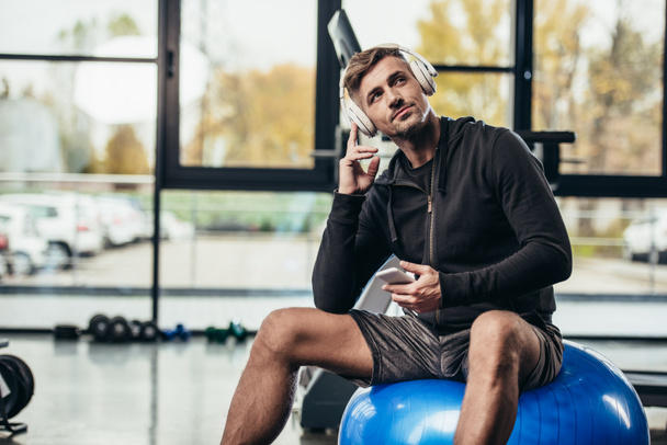 handsome sportsman sitting on fitness ball and listening music with smartphone in gym  - Zdjęcie, obraz