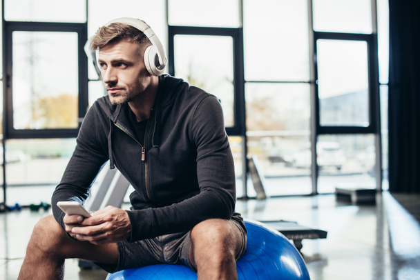 apuesto deportista sentado en la pelota de fitness con teléfono inteligente en el gimnasio
  - Foto, Imagen