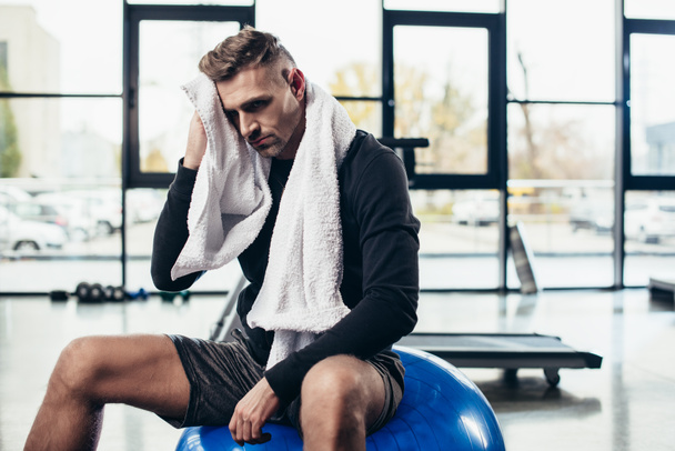handsome tired sportsman resting on fitness ball with towel in gym  - Zdjęcie, obraz