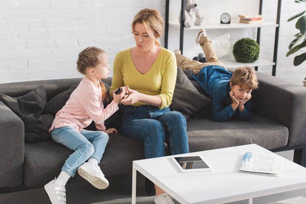 mother and daughter holding remote controller while boy lying on couch at home - Valokuva, kuva