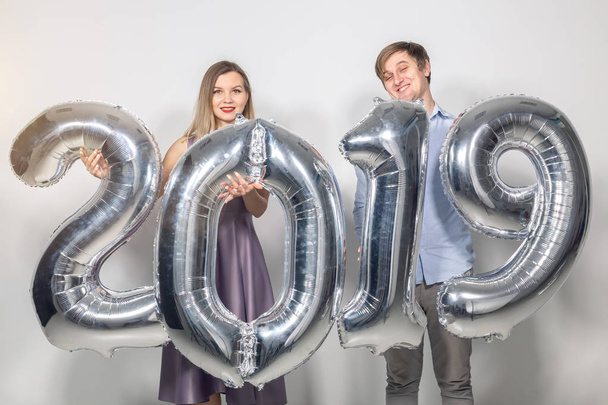 Πρωτοχρονιά, γιορτή και έννοιας διακοπές - αστεία αστεία αγάπη ζευγάρι κρατώντας υπογράφουν 2019 κατασκευασμένα από ασήμι μπαλόνια για το νέο έτος σε λευκό φόντο - Φωτογραφία, εικόνα